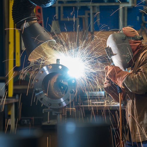 In einer Werkstatt schweißt ein Handwerker mit Schutzkleidung, während leuchtende Funken und Rauch um ihn herumwirbeln.