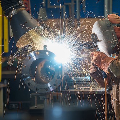 In einer Werkstatt schweißt ein Handwerker mit Schutzkleidung, während leuchtende Funken und Rauch um ihn herumwirbeln.