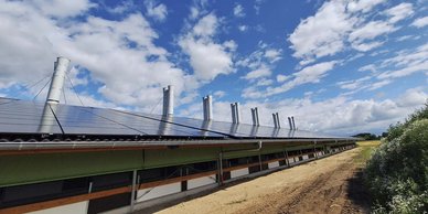 Ein landwirtschaftliches Gebäude mit mehreren Schornsteinen und Solarpanelen, unter einem strahlend blauen Himmel mit Wolken.