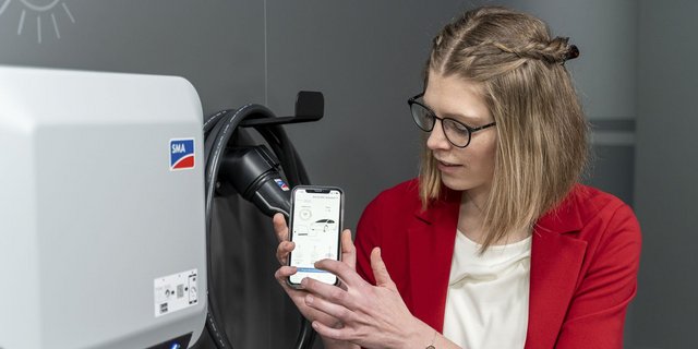 Eine Frau mit Brille zeigt auf ihr Smartphone, während sie neben einem modernen Ladegerät für Elektroautos steht.