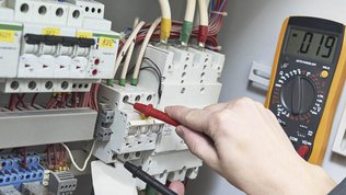 Das Bild zeigt eine Person, die mit einem Multimeter elektrische Verbindungen in einem Schaltkasten überprüft.
