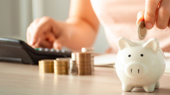 Auf einem Tisch wird eine Münze in ein weißes Sparschwein gesteckt, umgeben von Münzstapeln und einem Taschenrechner.