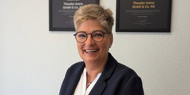 In einem hellen Büro lächelt eine Frau in einem Blazer, während sie vor gerahmten Zertifikaten steht, die an der Wand hängen.
